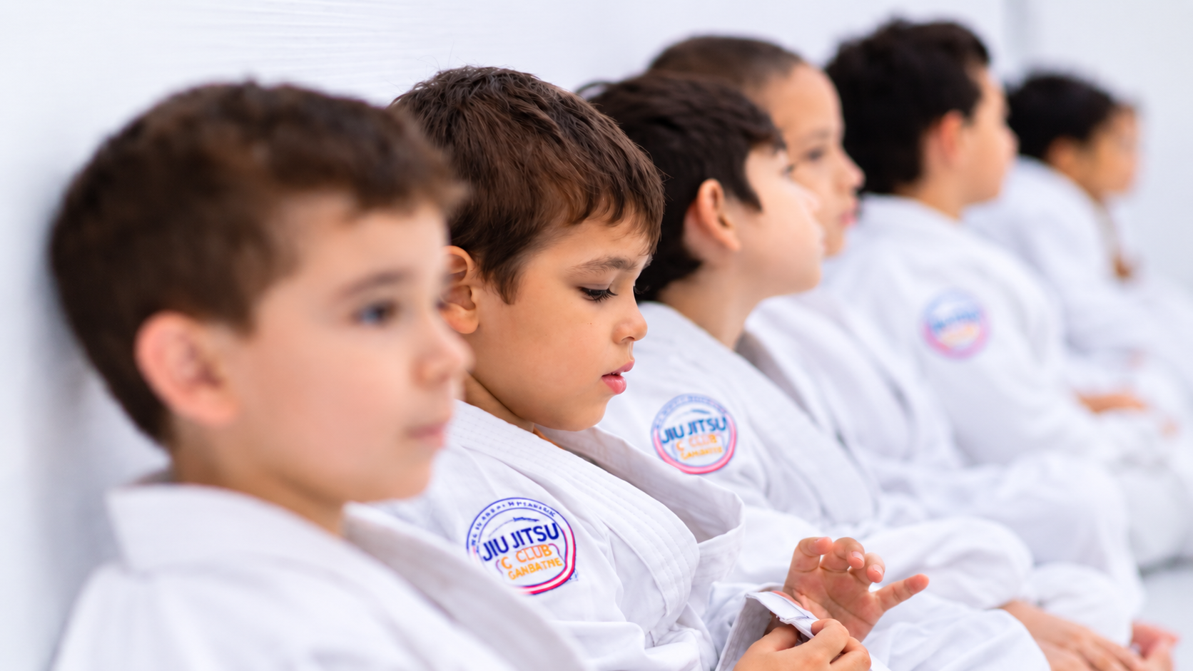 Crianças de kimono branco sentadas lado a lado durante aula de Jiu Jitsu, com o menino em primeiro plano em foco