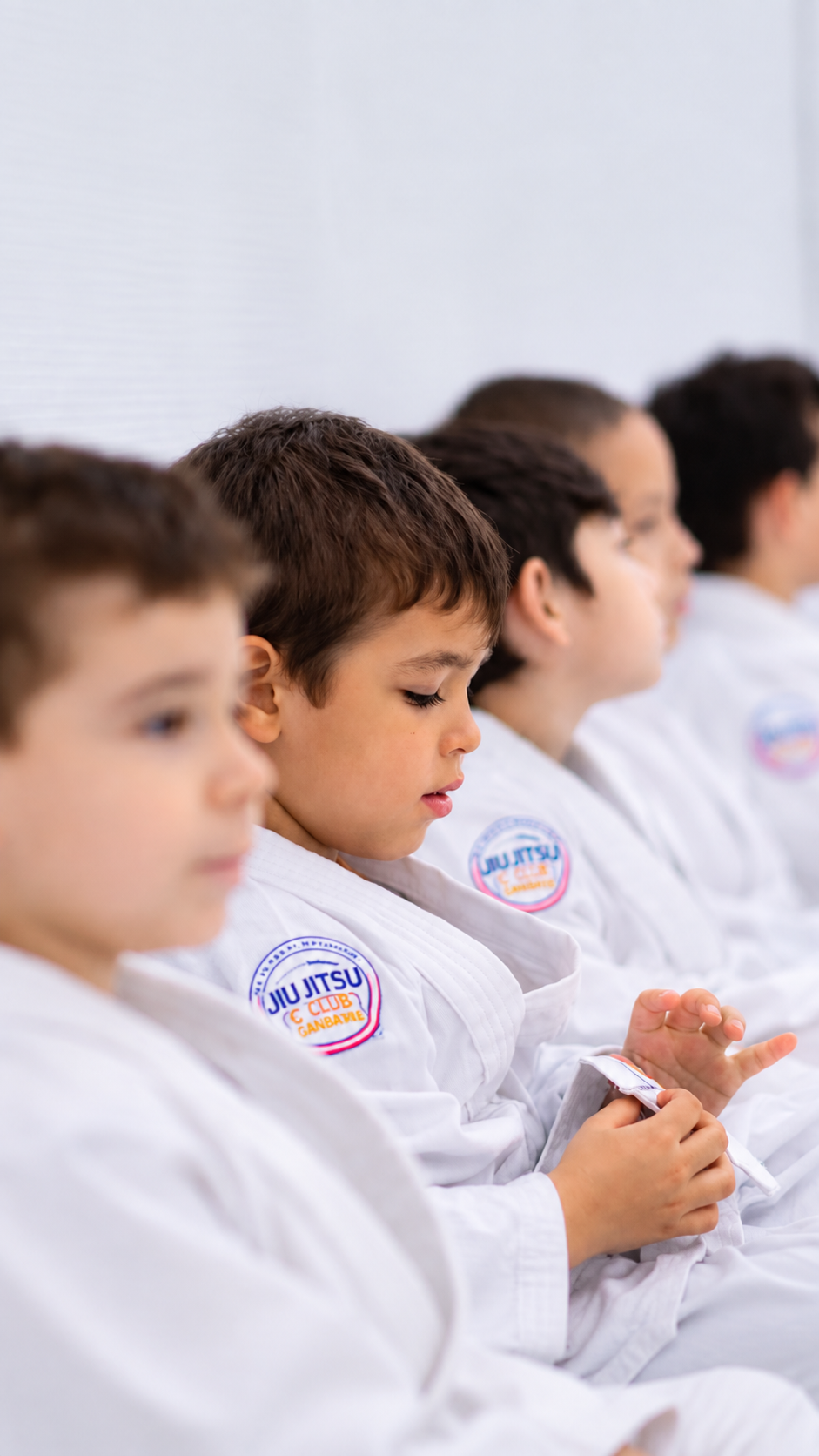 Menino de kimono branco sentado durante aula de Jiu Jitsu, com outras crianças ao fundo